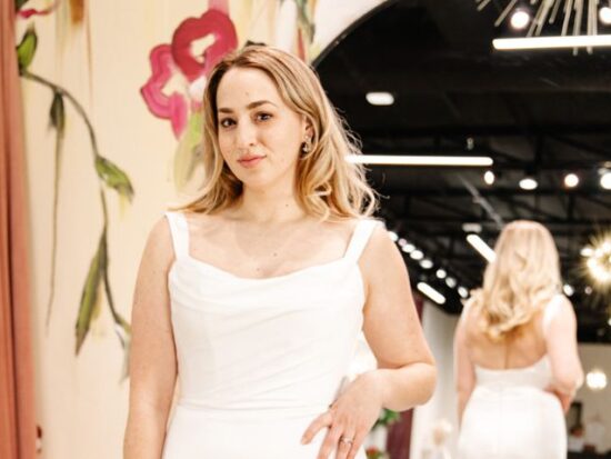 Bride in a square neckline wedding gown, standing in front of a floral mural with her reflection visible in a large round mirror.