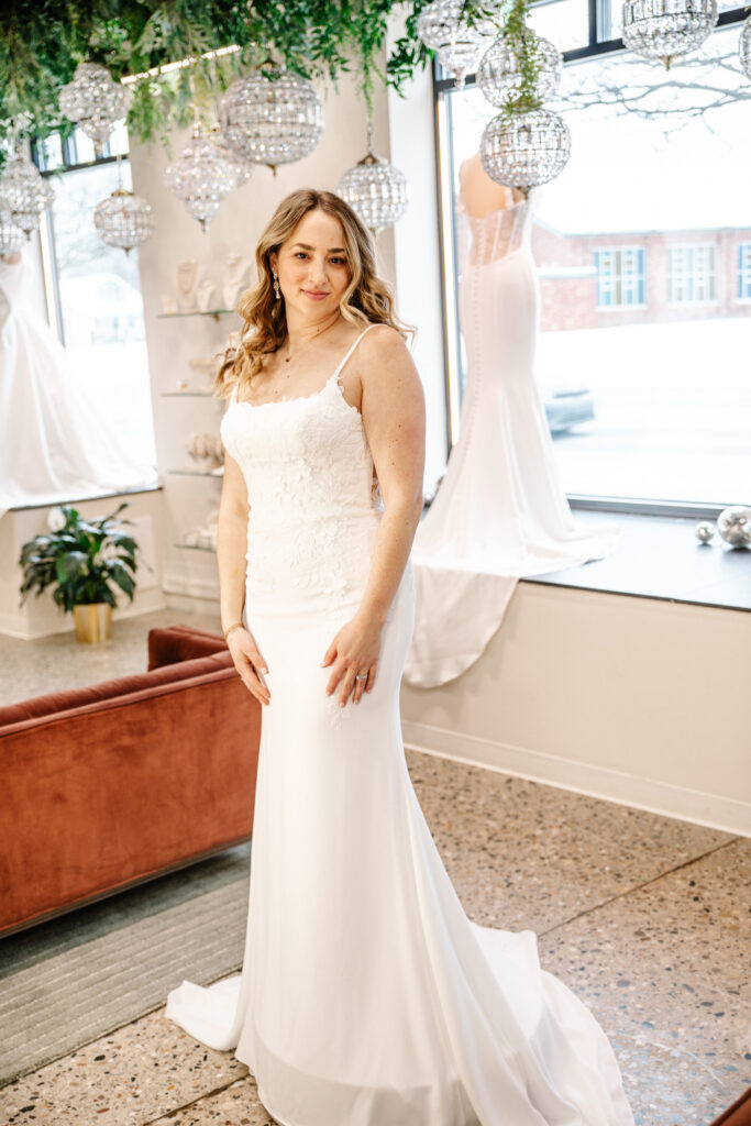 Bride in a fitted white gown with thin straps and floral appliqué, standing near large windows and crystal chandeliers.