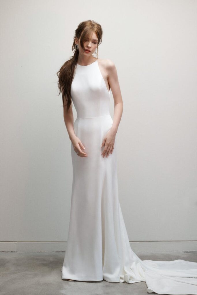 A bride models a modern high-neck, low-back wedding dress with a clean, sleek design and flowing train, posing in front of a soft gray wall.