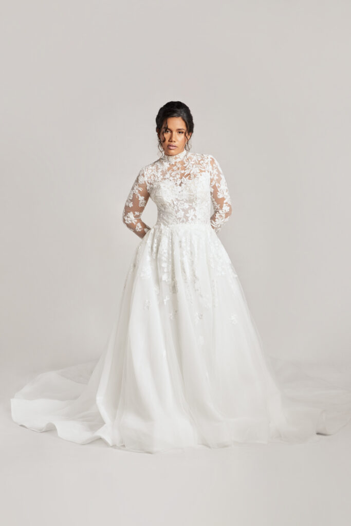 A bride stands confidently in a romantic high-neck long-sleeve wedding dress with allover lace detailing and a full tulle skirt, set against a light gray backdrop.