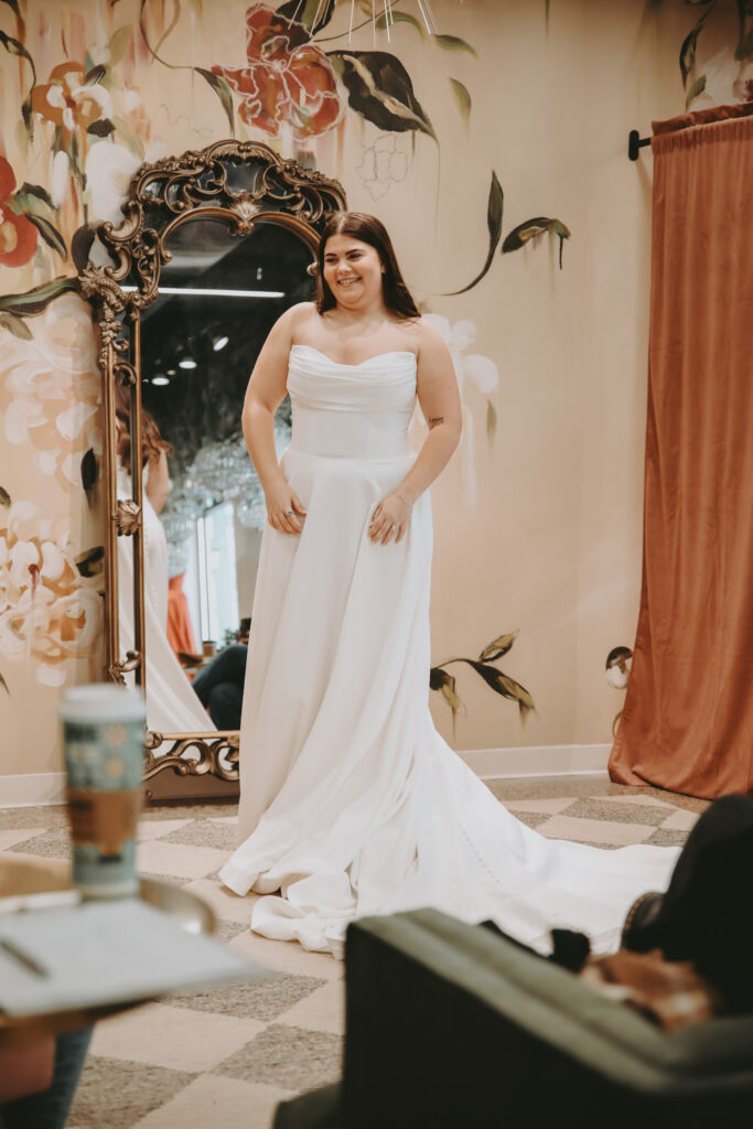 A smiling bride stands in front of an ornate mirror wearing a strapless satin wedding gown with ruching on the bodice and a flowing train.