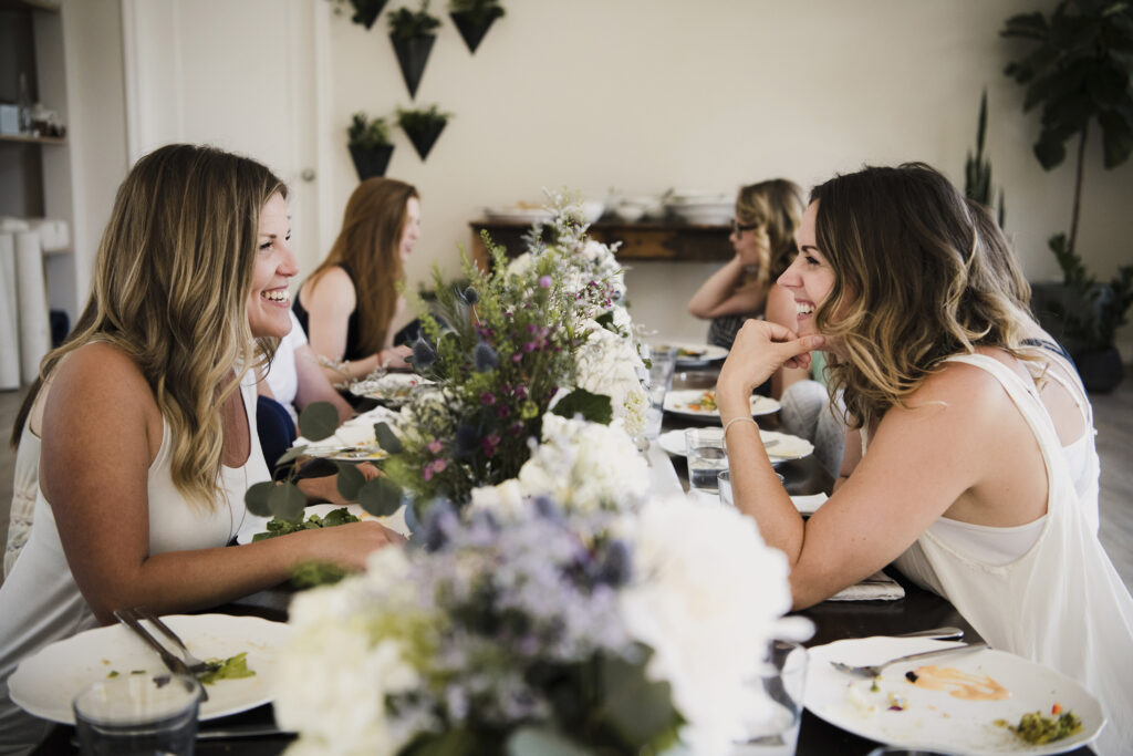 Two women seated at a beautifully decorated table smiling and chatting during a daytime bridal shower celebration with floral centerpieces.