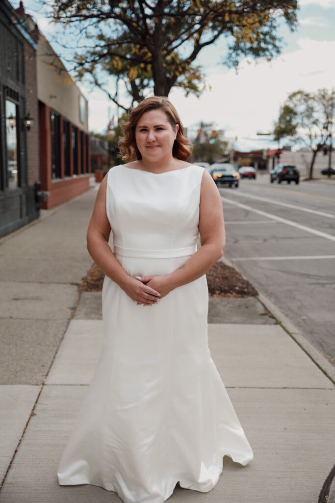 A bride stands on a city sidewalk wearing a classic satin wedding dress with a high bateau neckline and waistband, giving a timeless and elegant look.