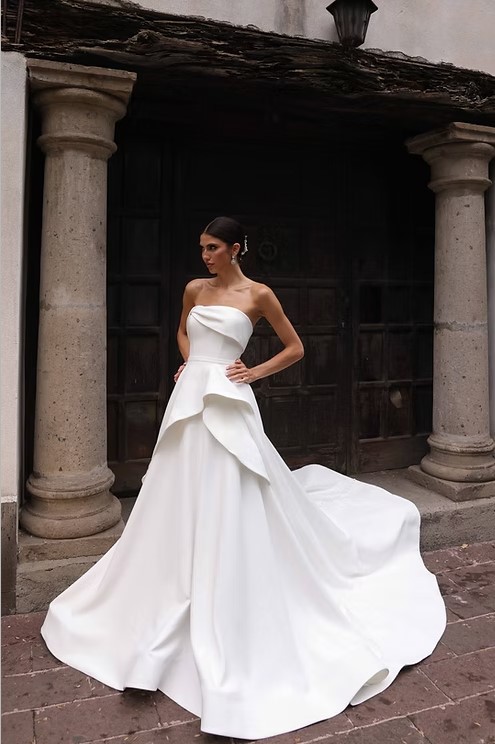 A bride in a strapless wedding dress with an architectural overskirt and dramatic train poses outside near stone columns and a wooden door.