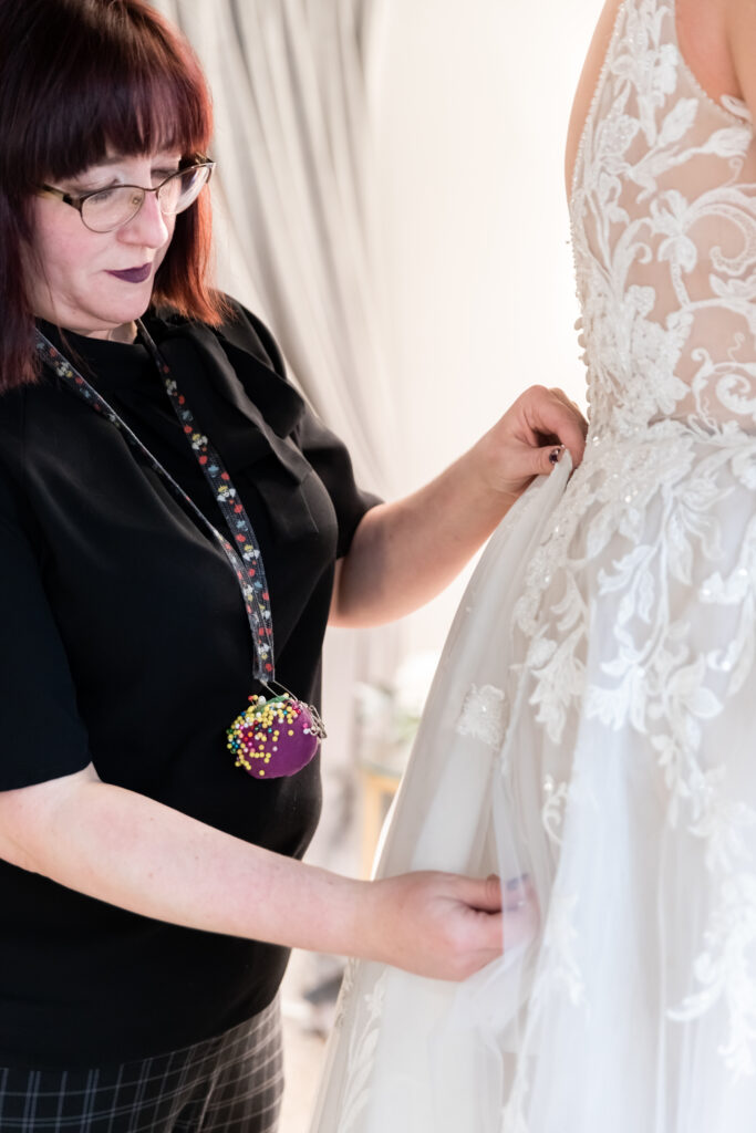 A seamstress adjusts the layers of a lace wedding dress during a bridal gown fitting session.