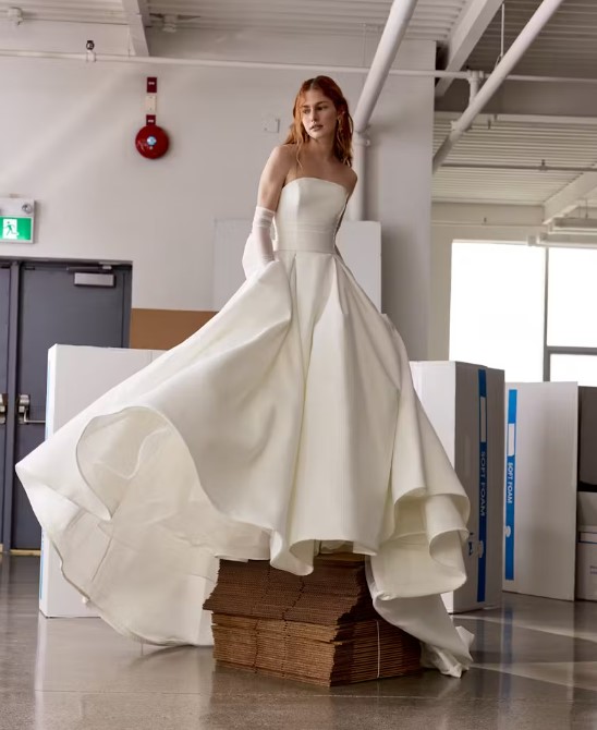 A bride standing on stacked cardboard wearing a strapless ballgown with a dramatic skirt, surrounded by a warehouse-like interior.