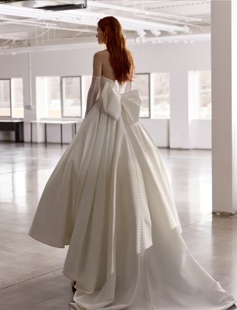 Rear view of a bride in a strapless gown with a large bow and trailing buttons down the back, standing in a bright, open room.