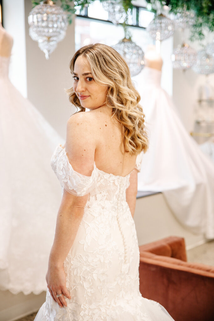 A bride looks over her shoulder in an off-the-shoulder lace wedding dress with crystal chandeliers behind her.