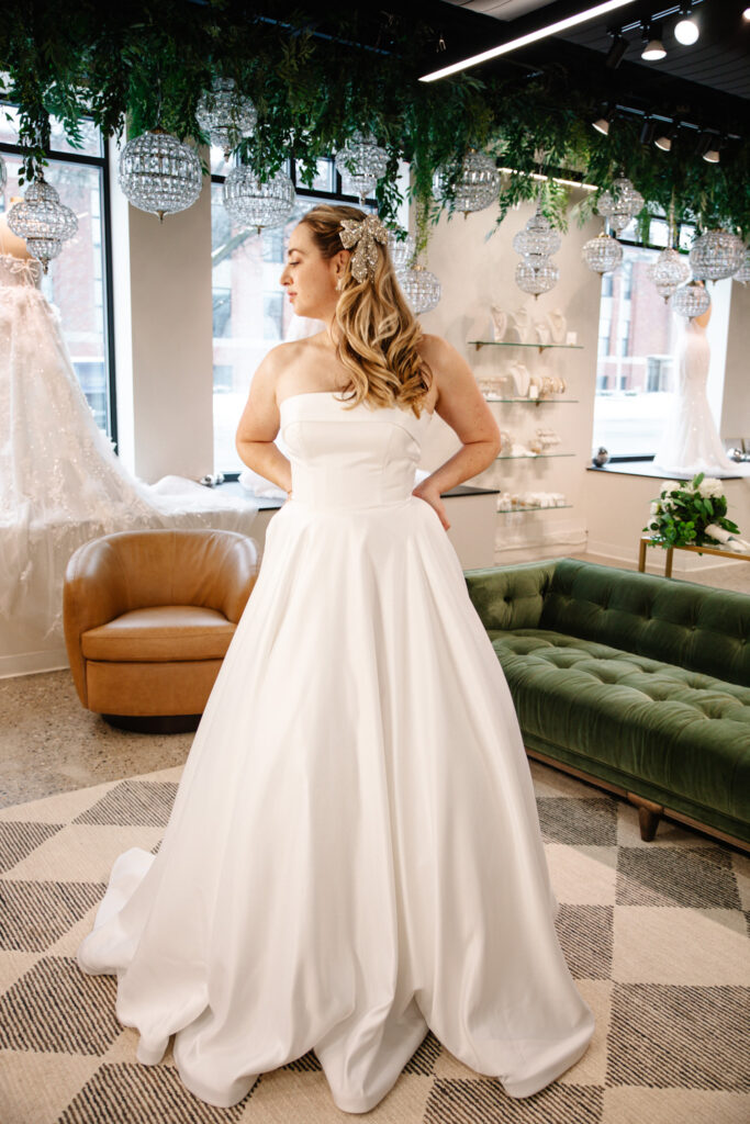 A bride with curled hair and a sparkly bow clip poses in a strapless ballgown inside a bridal shop with chandeliers and greenery.