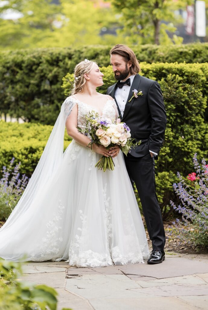 Bride and groom standing outdoors, bride in a lace wedding gown with a floral bouquet, gazing at each other.