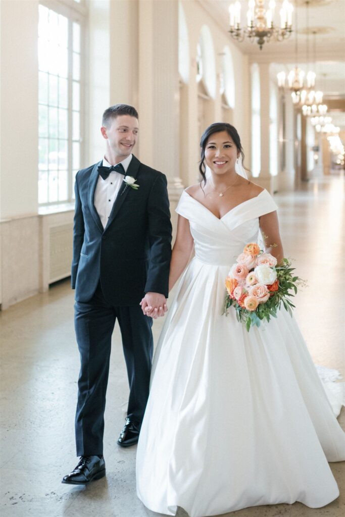 Bride and groom walking hand-in-hand through a grand hallway, bride wearing a classic off-the-shoulder gown with a peach bouquet.