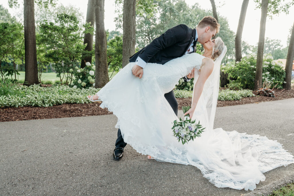 Groom dips and kisses bride in lace wedding dress on a tree-lined path, bride holding a cascading bouquet.