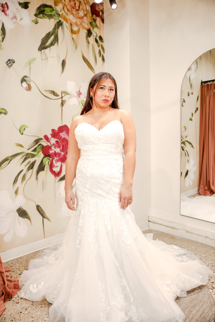 Bride in a sweetheart lace mermaid wedding gown with a tulle train, standing against a painted floral wall and looking forward.