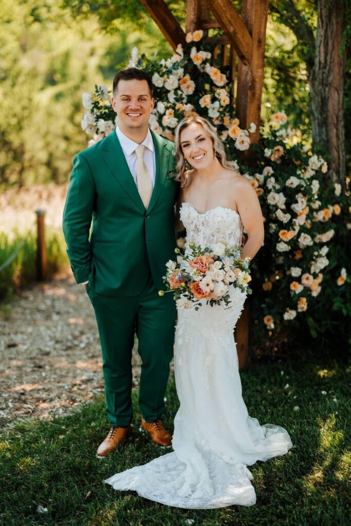 Bride and groom smiling under a wooden arch adorned with flowers, bride in a strapless gown with a peach and cream bouquet.
