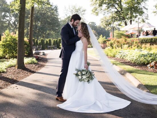 Bride and groom in garden setting, bride in a floral lace dress holding a pastel bouquet, groom in a black tuxedo.