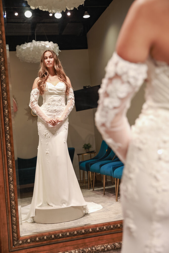 A bride stands in front of a mirror wearing a fitted gown with long sheer lace sleeves and floral embroidery.