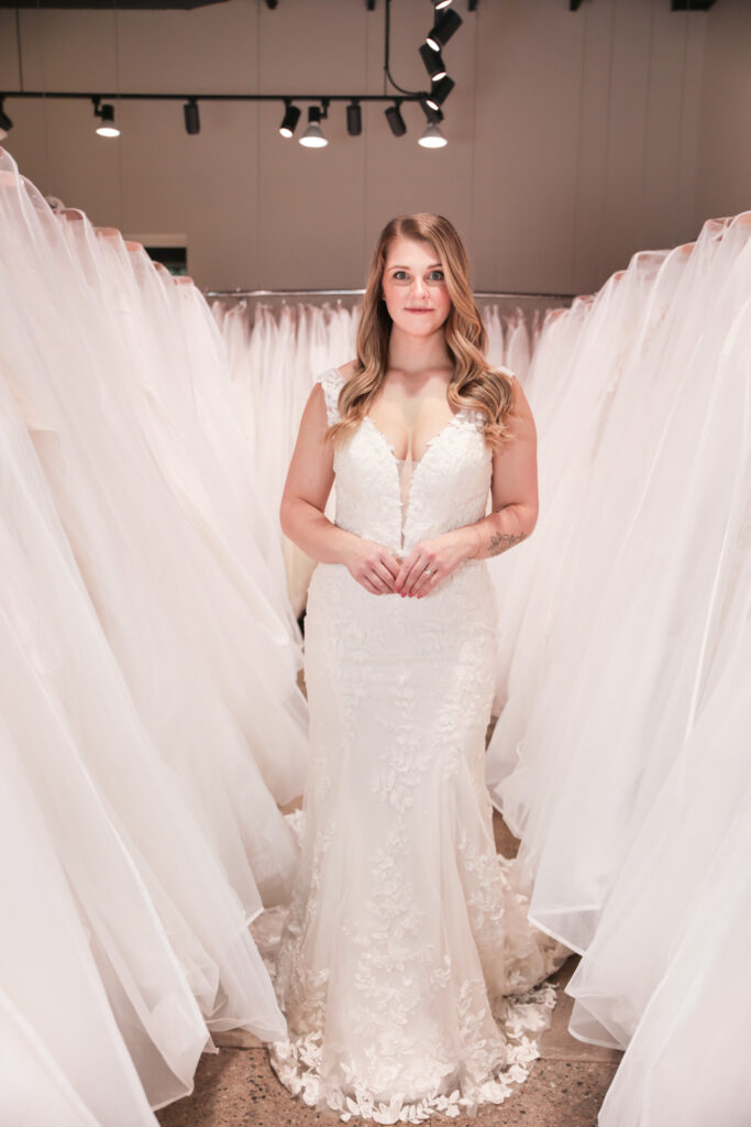Bride standing between racks of gowns, wearing a fitted lace wedding dress with a plunging neckline and cap sleeves.