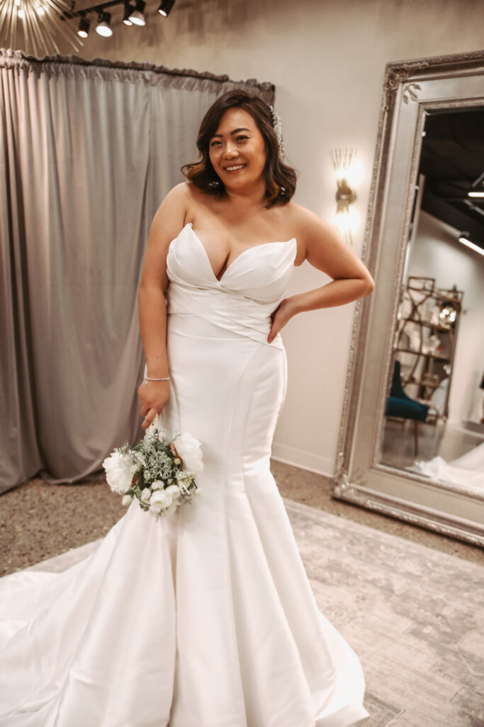 Bride smiling in a strapless satin fit-and-flare wedding dress, holding a white bouquet in a bridal fitting room.