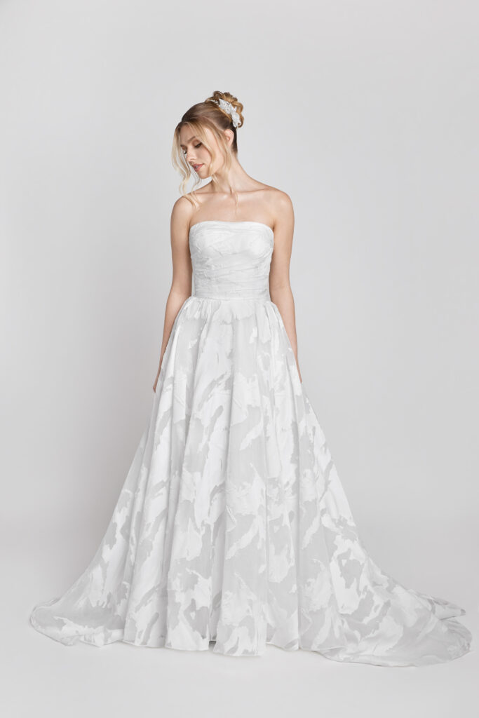 A bride in a textured strapless A-line wedding dress with soft floral patterns looks down, standing against a light gray background.