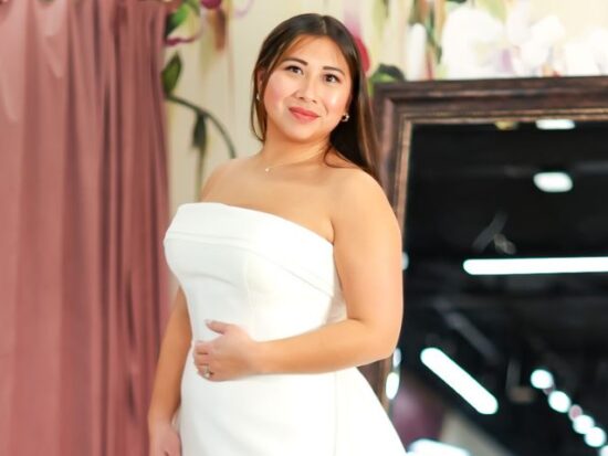 A bride poses in a strapless structured wedding gown in front of mauve curtains and a full-length mirror at a bridal boutique.