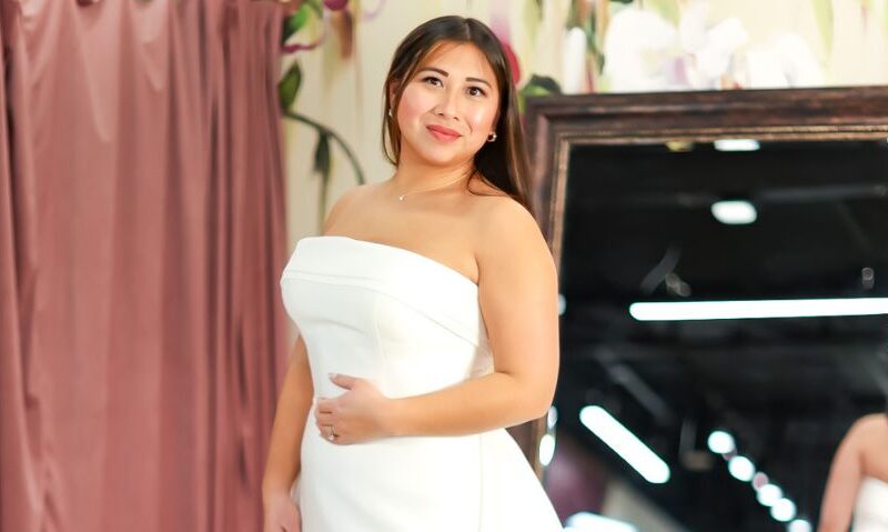 A bride poses in a strapless structured wedding gown in front of mauve curtains and a full-length mirror at a bridal boutique.