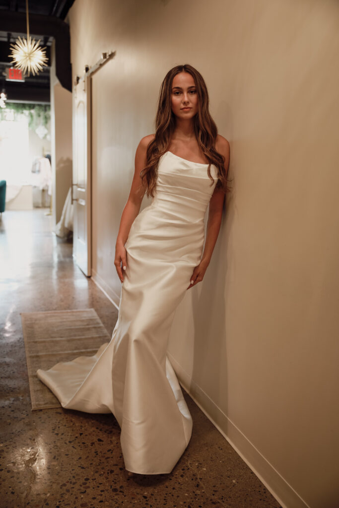 A bride in a one-shoulder satin fit-and-flare gown leans against a hallway wall, showcasing the sleek silhouette and train of the dress.