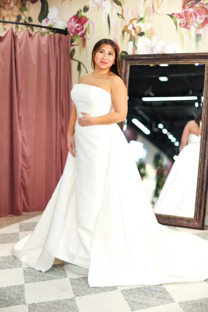 A full-length view of a bride wearing a sleek strapless wedding dress with clean lines and a structured silhouette, posing confidently.