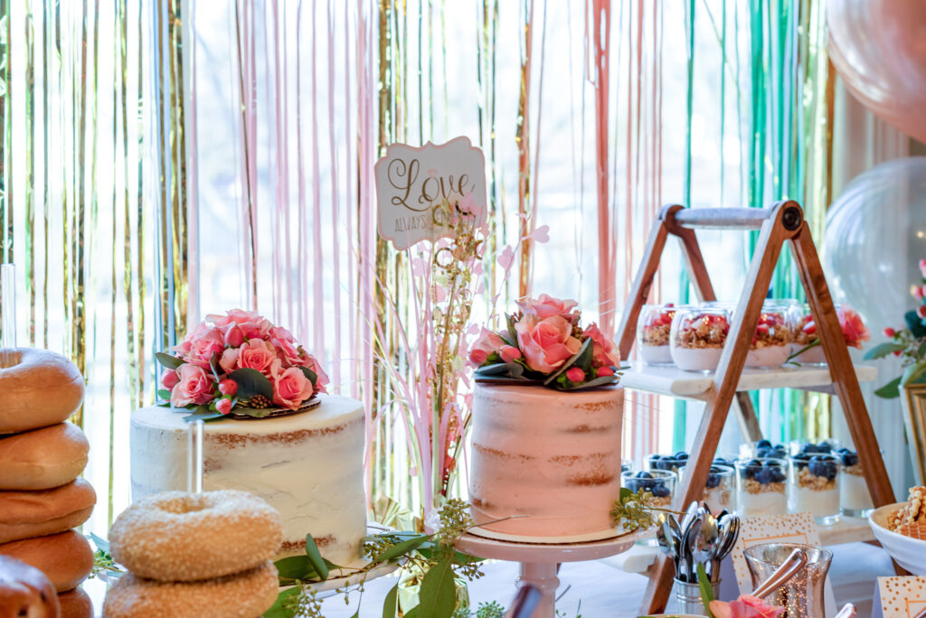Two-tiered wedding shower cakes decorated with pink rose toppers and surrounded by yogurt parfaits and bagels on a colorful dessert table.