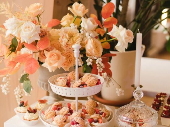 Tabletop filled with floral decor and sweets, including a two-tiered dessert tray with cream puffs and berries, surrounded by orange and ivory flowers.