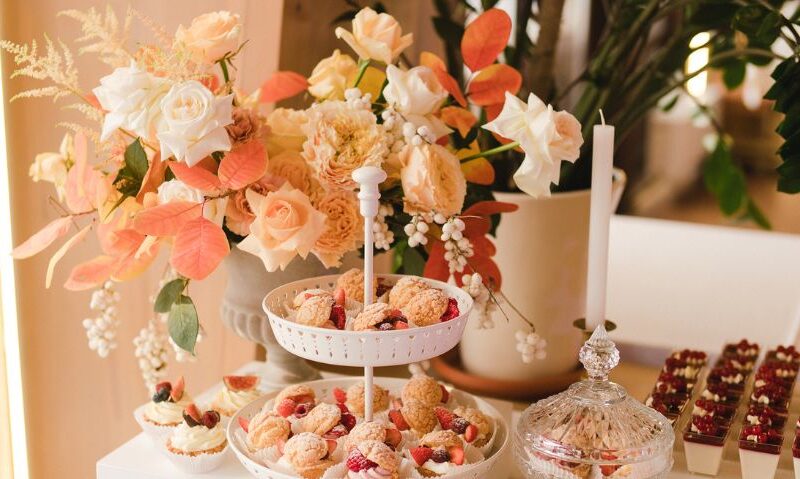 Tabletop filled with floral decor and sweets, including a two-tiered dessert tray with cream puffs and berries, surrounded by orange and ivory flowers.