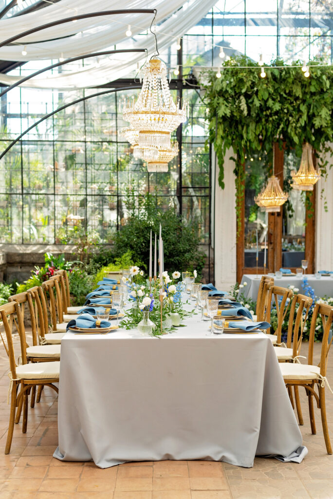 Stylish wedding shower table with blue and white floral centerpieces, tall candles, and wooden chairs set under hanging chandeliers.