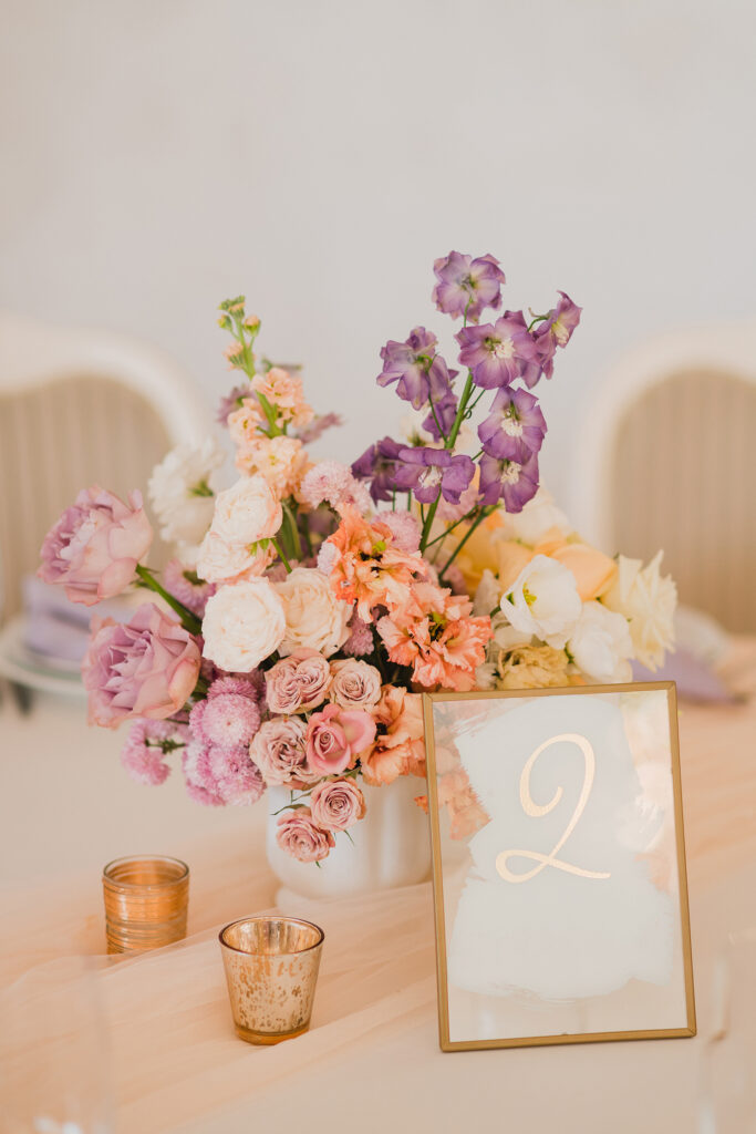 Close-up of pastel-toned flower arrangement with roses, peonies, and lisianthus beside a gold-framed table number and votive candles.