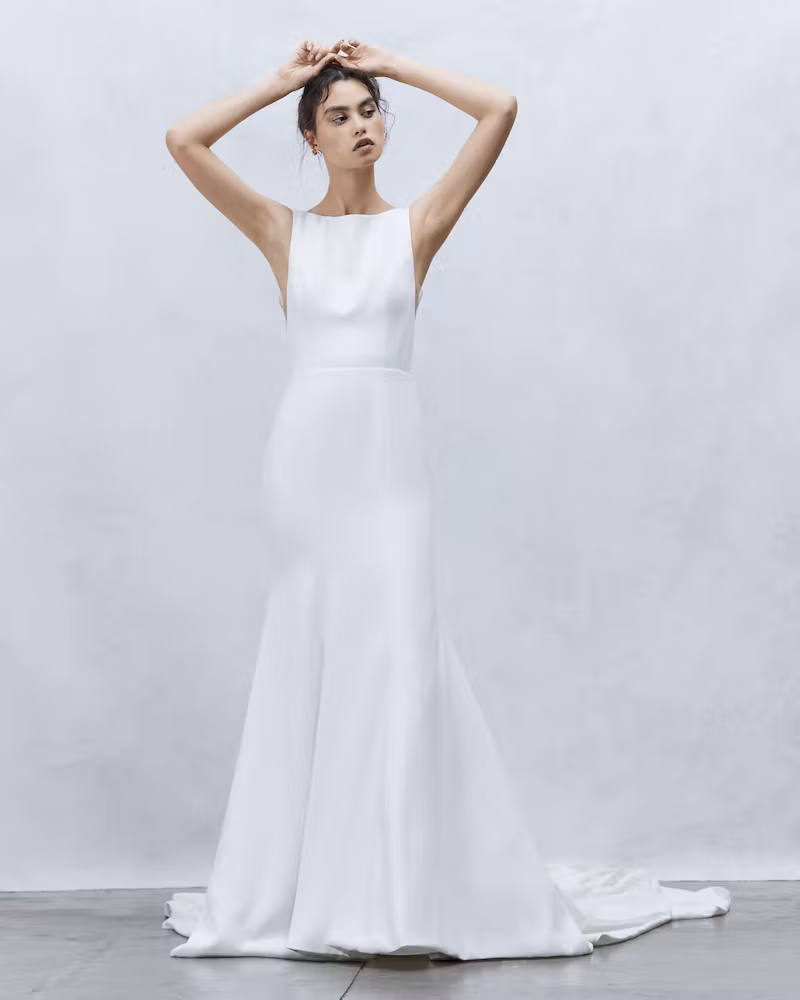 Bride in a modest wedding dress standing with her hands resting on her head in front of a plain white background.