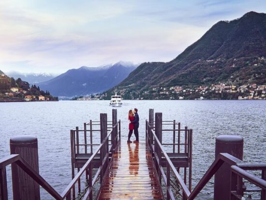 A couple embraces at the end of a wooden dock, surrounded by a peaceful lake with a boat in the distance and mountain villages on either side.
