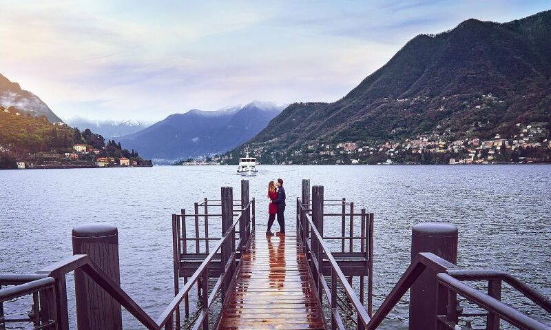A couple embraces at the end of a wooden dock, surrounded by a peaceful lake with a boat in the distance and mountain villages on either side.