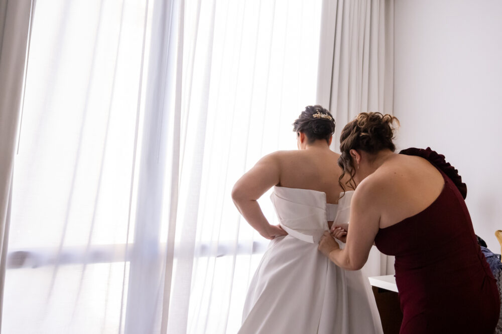Woman in burgundy dress helping bride into a strapless satin wedding gown by a sheer curtain.
