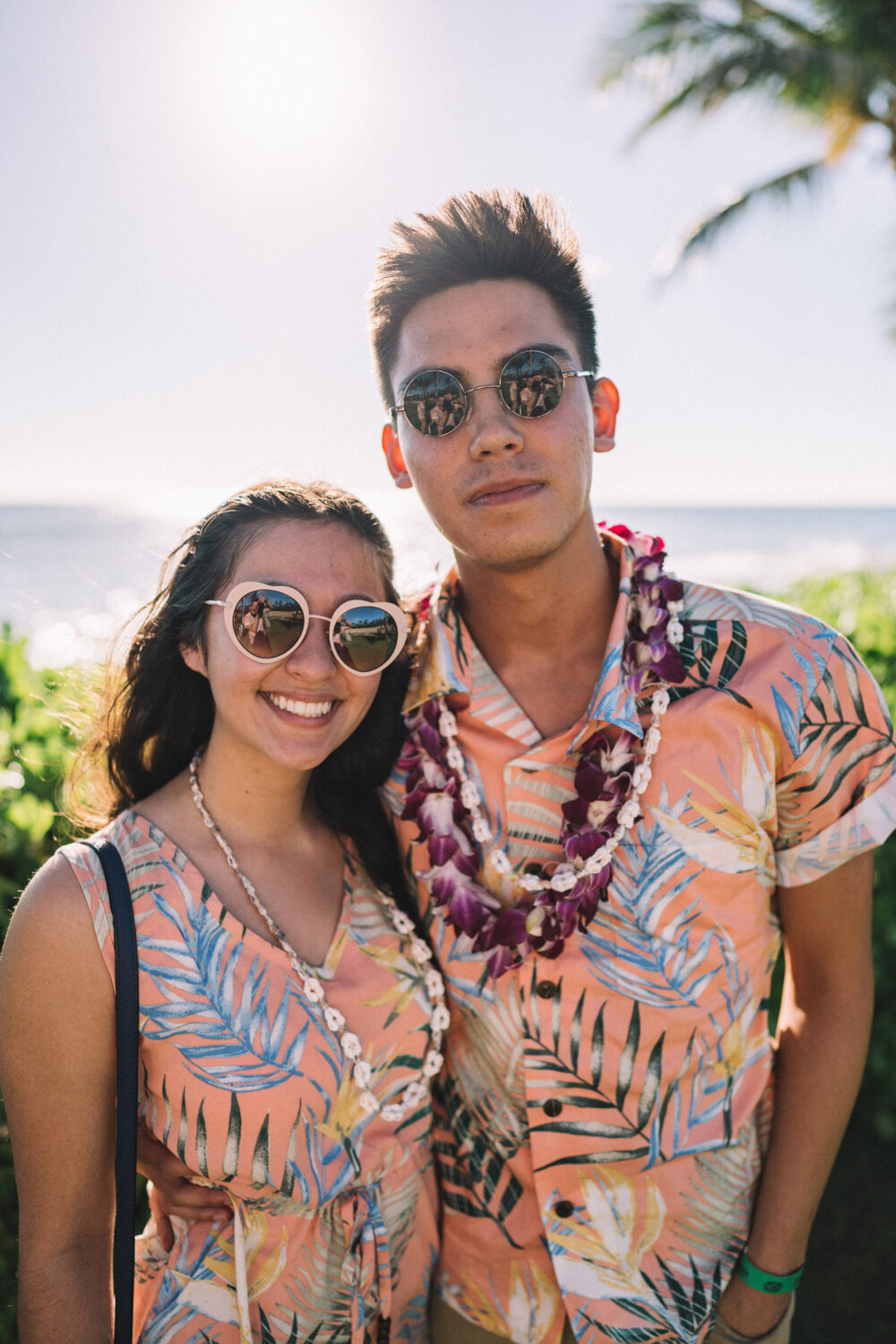 A smiling couple poses in matching tropical outfits and sunglasses at a beachside location, wearing leis and soaking in the sunlight.