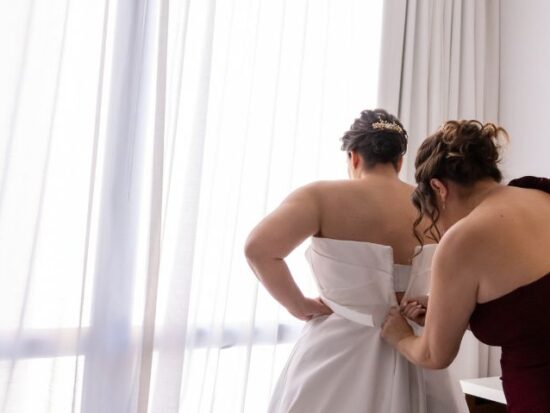 Woman in burgundy dress helping bride into a strapless satin wedding gown by a sheer curtain.