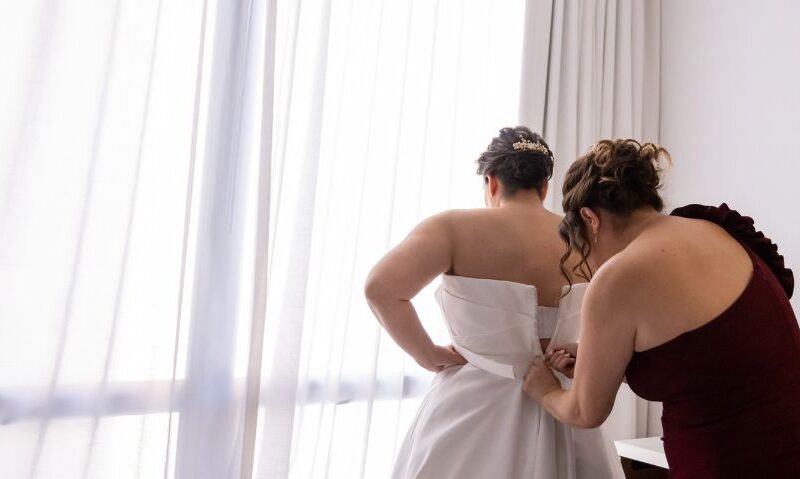 Woman in burgundy dress helping bride into a strapless satin wedding gown by a sheer curtain.