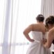 Woman in burgundy dress helping bride into a strapless satin wedding gown by a sheer curtain.
