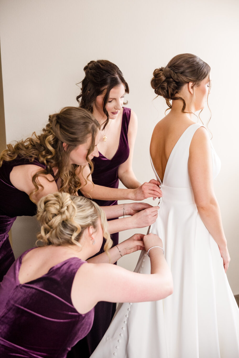Bridesmaids in purple dresses buttoning bride’s satin gown with a deep V-back.