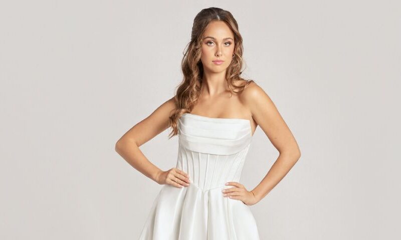 A studio image of a model wearing a strapless white corset wedding dress with a pleated satin bodice and flowing ballgown silhouette.