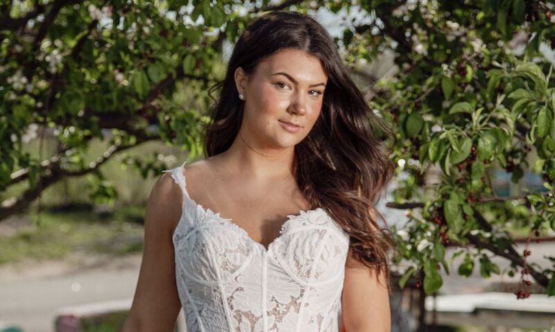A bride stands outdoors in front of blossoming trees wearing a short lace wedding dress with a structured bodice and sweetheart neckline.
