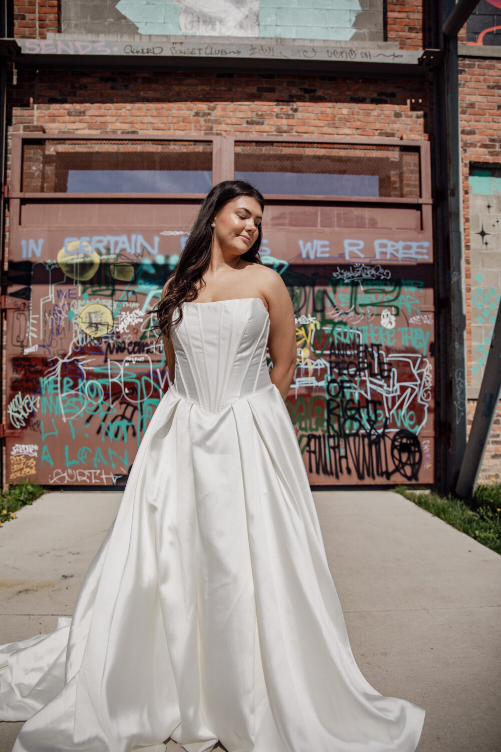 A bride stands outside in front of a graffiti-covered wall wearing a strapless satin ballgown with a pleated skirt and structured corset bodice.