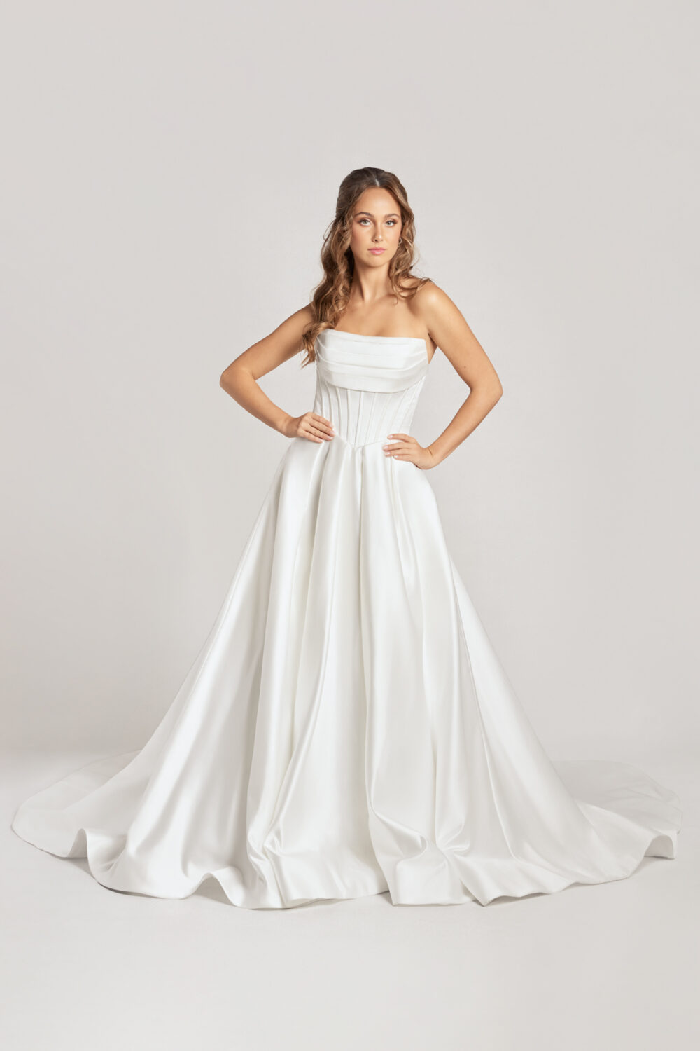 A model poses in a studio wearing a strapless satin wedding gown with a hand-draped corset bodice and a full, flowing ballgown skirt.