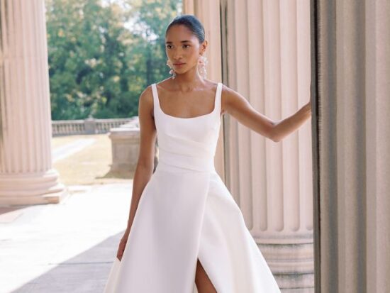 A bride stands confidently between tall columns wearing a modern tank-strap wedding gown with a high slit.