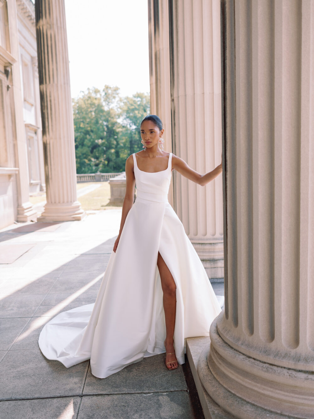 A bride stands confidently between tall columns wearing a modern tank-strap wedding gown with a high slit.