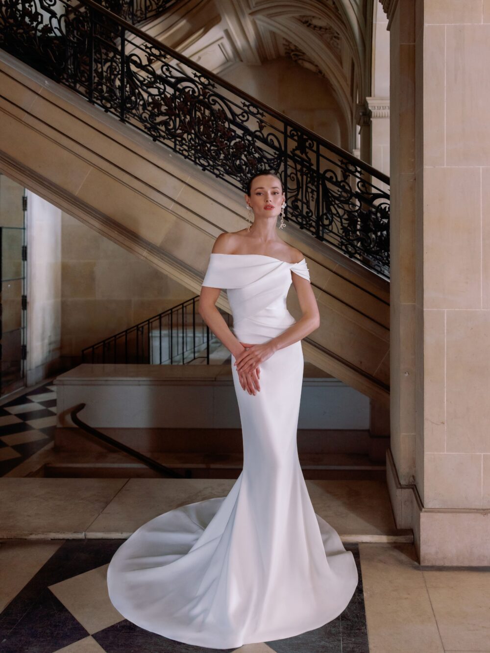 A bride poses near an elegant staircase wearing an off-the-shoulder satin Sareh Nouri gown with a fitted silhouette.