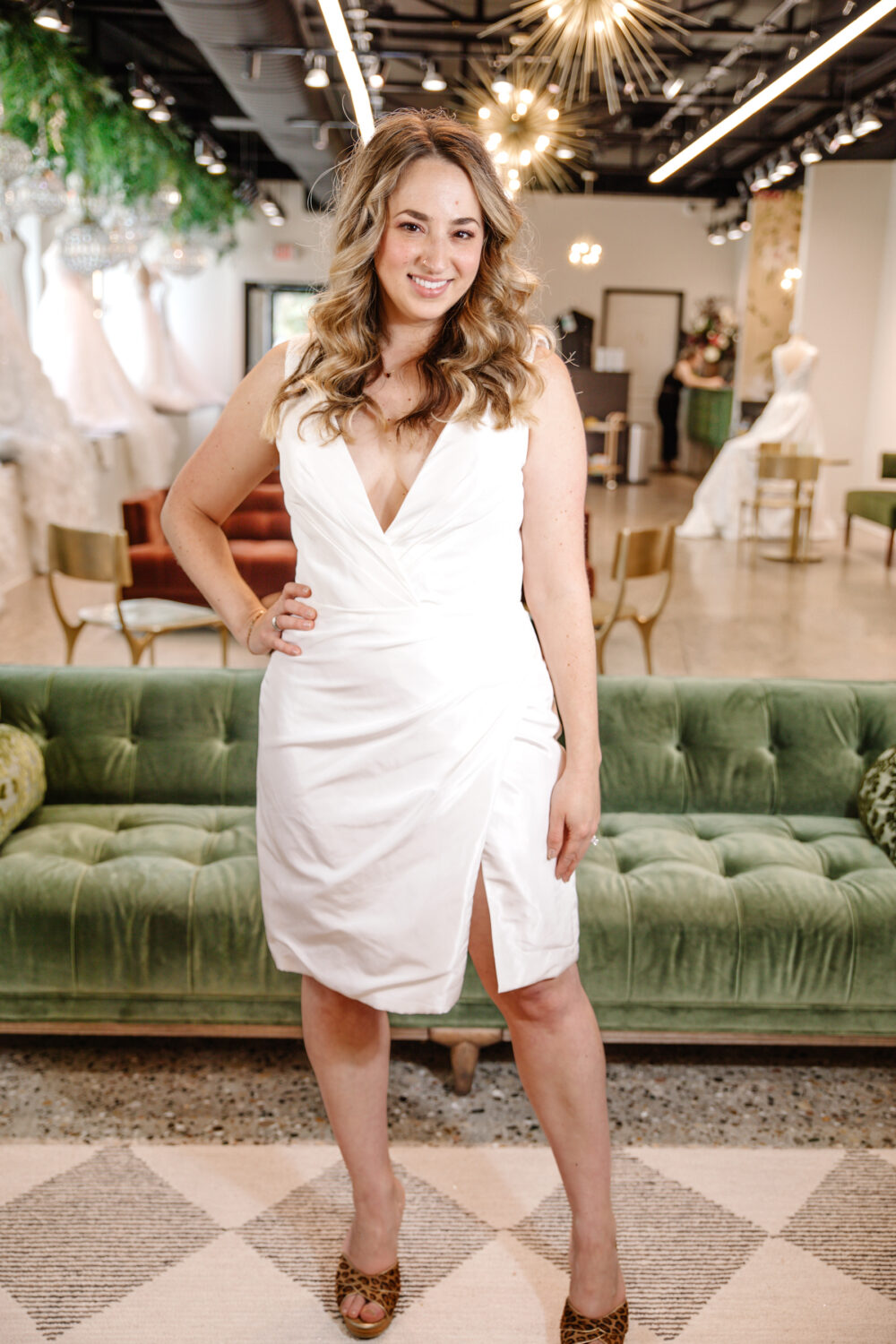 A bride stands confidently in a short white wrap-style wedding dress with a deep V-neckline and front slit, posing inside The Wedding Shoppe.