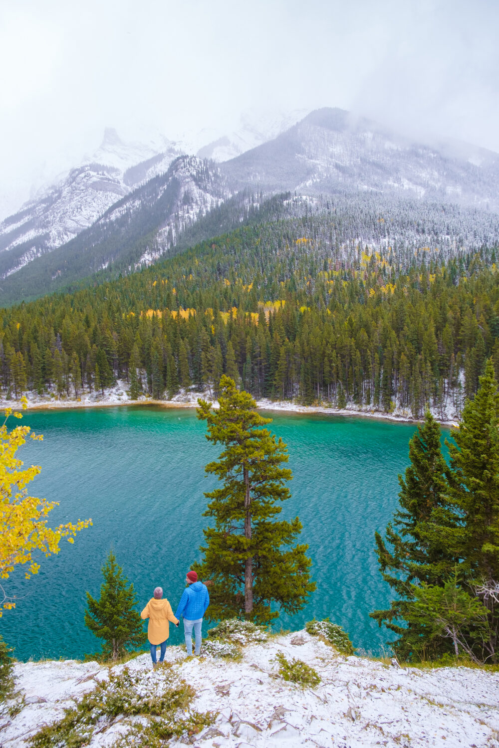 A couple stands hand in hand on a snowy cliffside, looking out over a turquoise lake and pine forest with misty mountains in the distance.